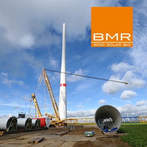 A wind turbine is being constructed, featuring cranes and turbine components in the foreground under a cloudy sky.