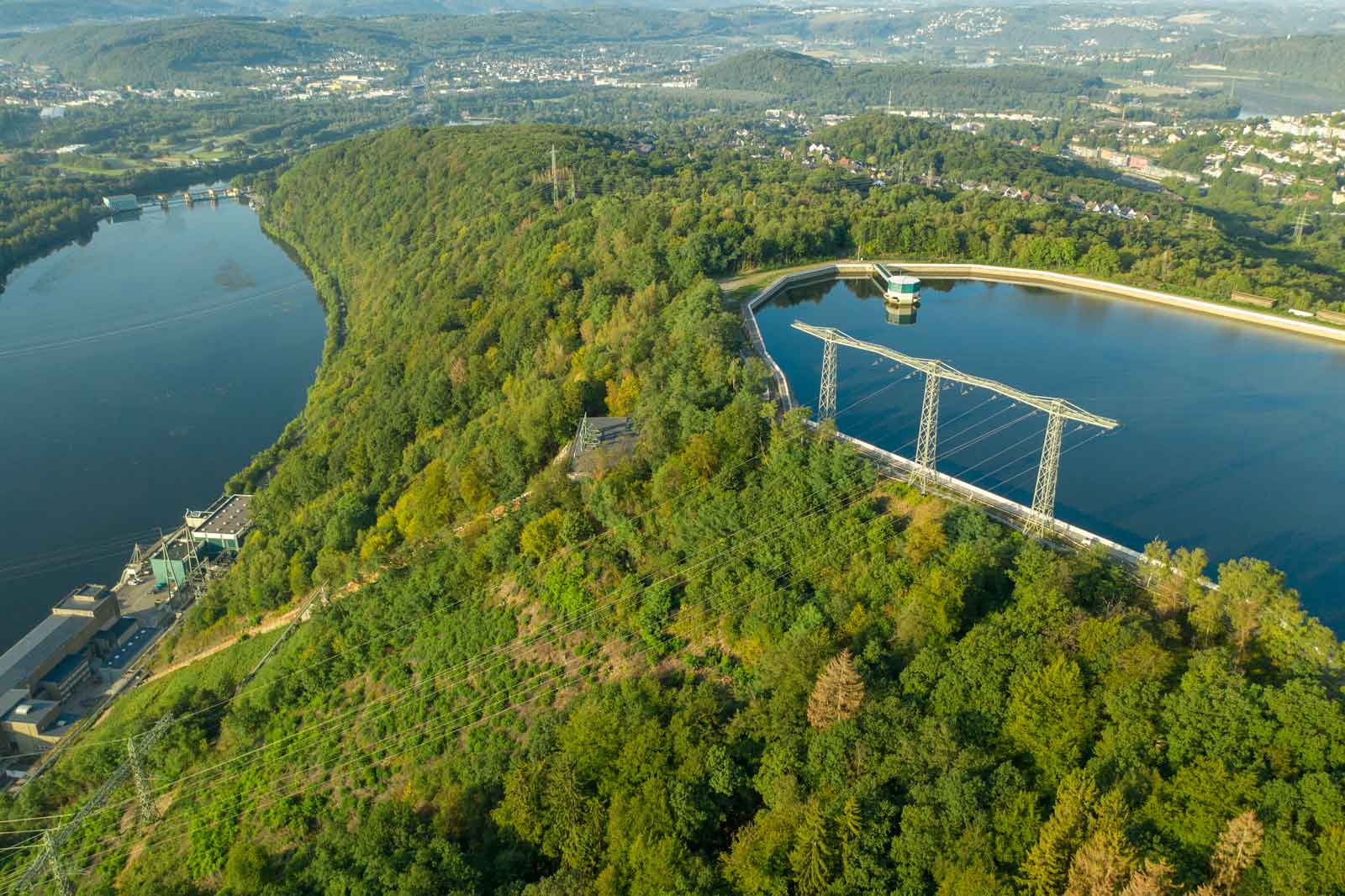 Eine Luftaufnahme eines bewaldeten Hügels neben einem See, mit elektrischen Masten und einem Wasserspeicher.