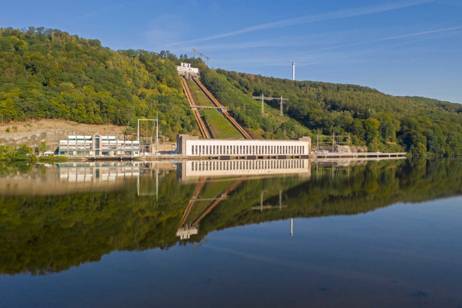 Ein Wasserkraftwerk am Ufer eines ruhigen Sees, umgeben von bewaldeten Hügeln, spiegelt sich im Wasser wider.