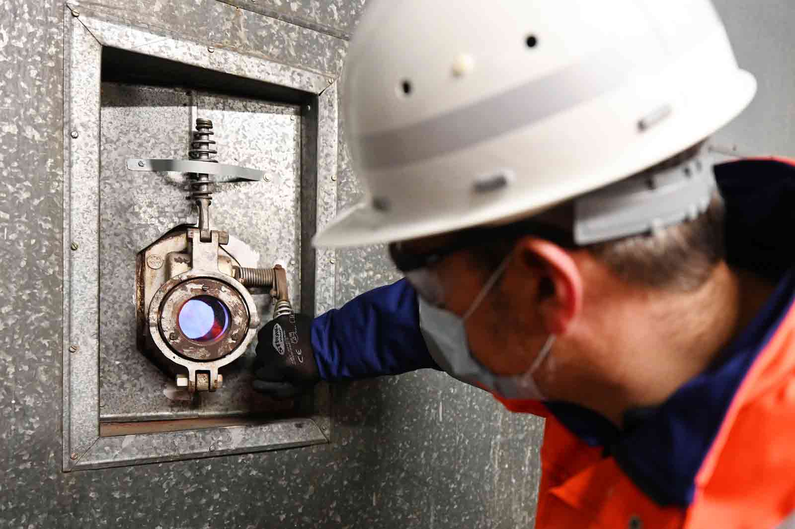 Ein Arbeiter in einem Schutzhelm und Handschuhen bedient ein technisches Gerät, das in einer metallischen Wand installiert ist.
