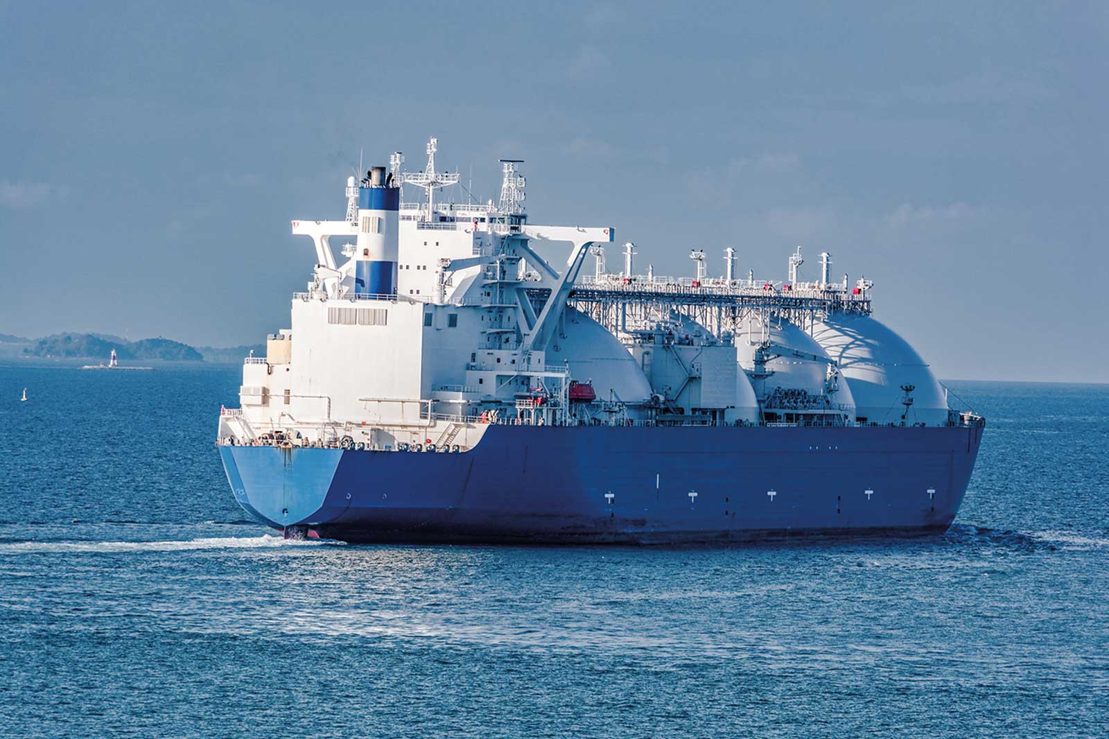 Ein Frachtschiff für Flüssiggas fährt über ruhiges Wasser, mit blauer Farbe und weißen Tanks, unter einem klaren Himmel.