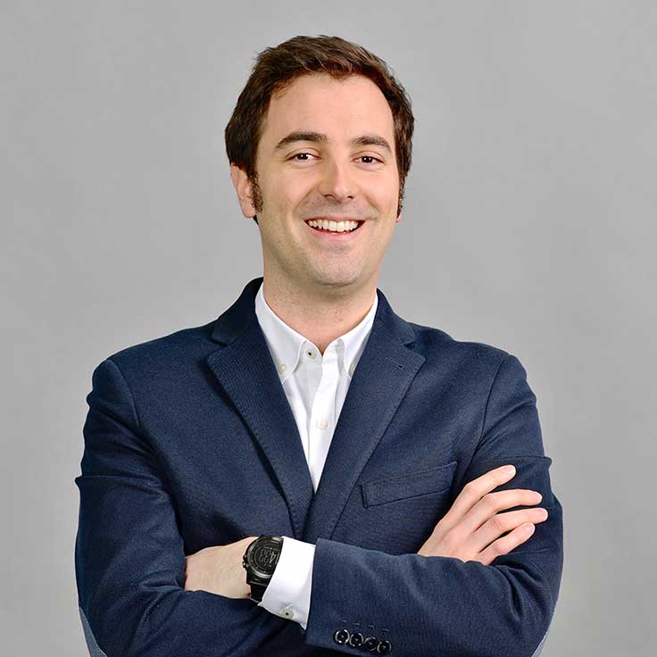 A man in a dark blazer stands with crossed arms, wearing a white shirt and a wristwatch, against a grey background.