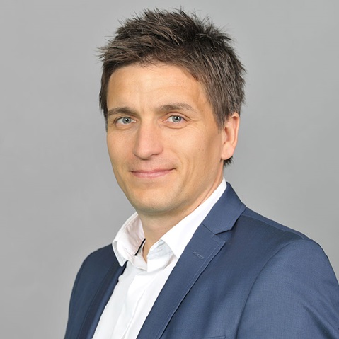 A man in a blue suit and white shirt stands against a grey background.