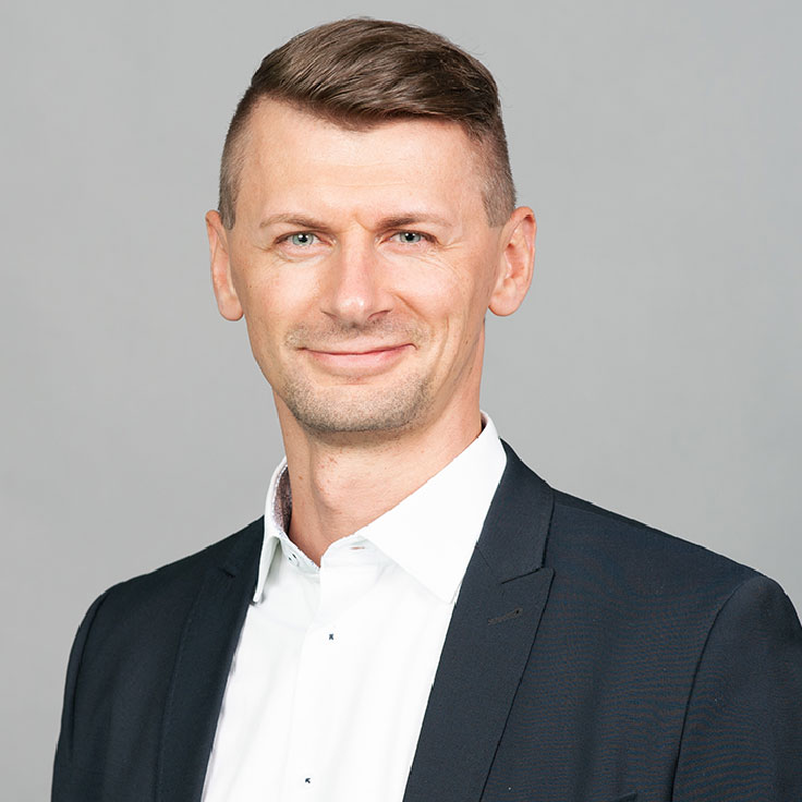 A man in a black suit and white shirt poses against a grey background, showcasing a professional appearance.