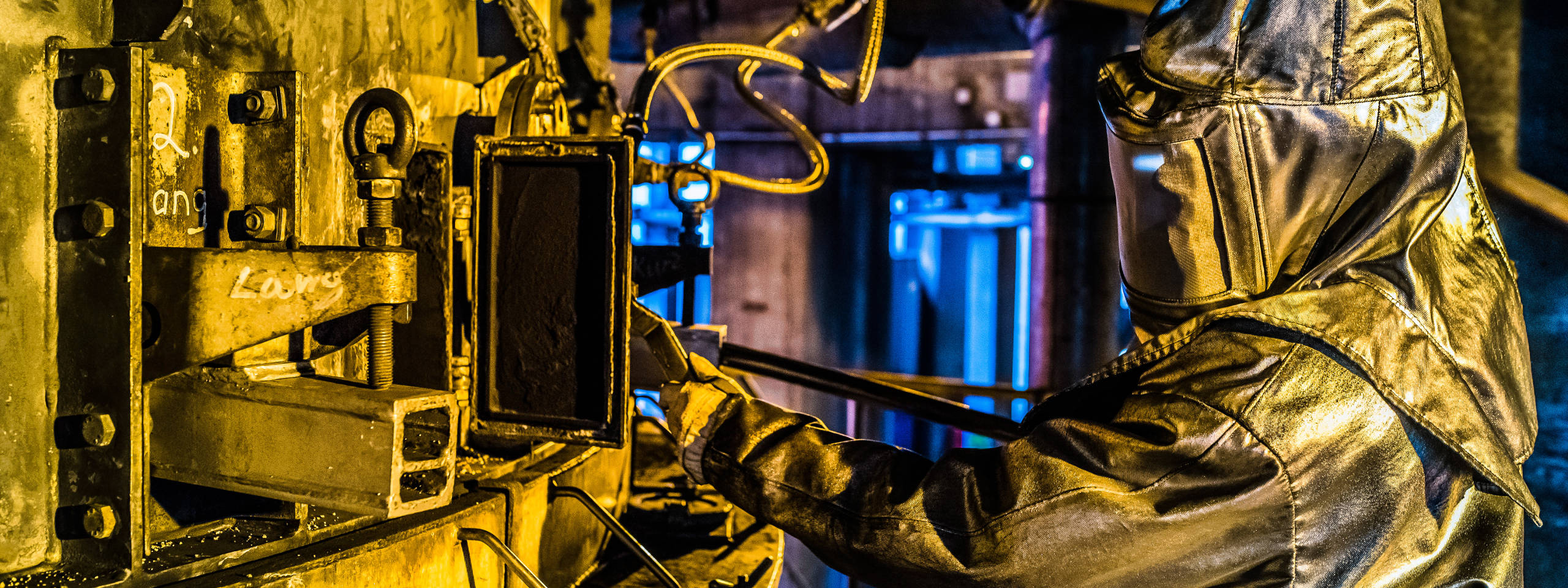 Ein Arbeiter trägt einen Schutzanzug und bedient Maschinen in einer industriellen Umgebung mit gelben und blauen Lichtern.