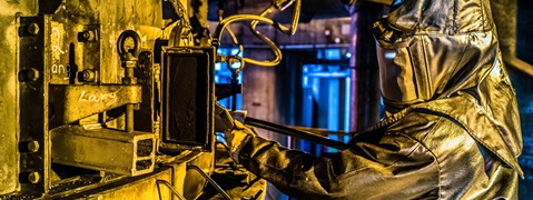 Ein Arbeiter trägt einen Schutzanzug und bedient Maschinen in einer industriellen Umgebung mit gelben und blauen Lichtern.