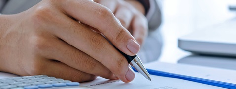 Eine Hand hält einen Stift über einem Dokument, neben einem Taschenrechner und einem Laptop.