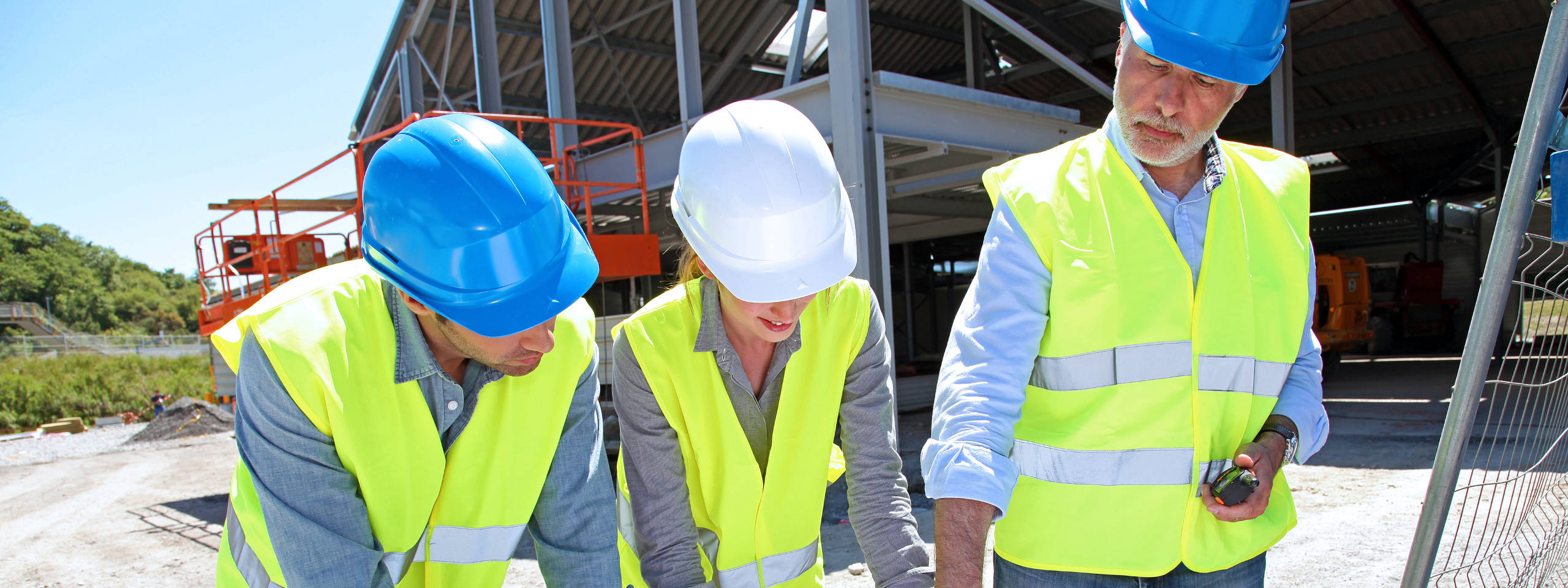 Three construction workers in yellow safety vests and helmets reviewing plans at a construction site under clear skies.