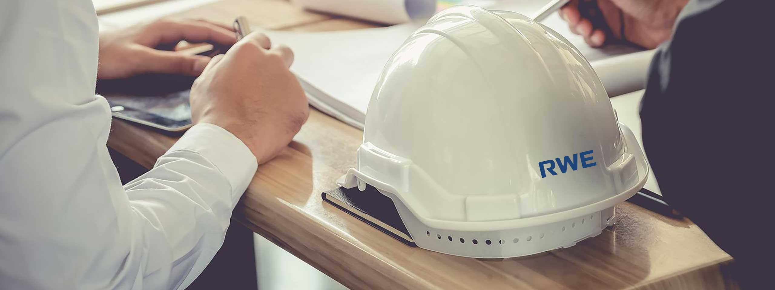 Ein weißer Helm mit dem Logo 'RWE' auf einem Holztisch, mit Händen, die schreiben, und einem Tablet im Hintergrund.