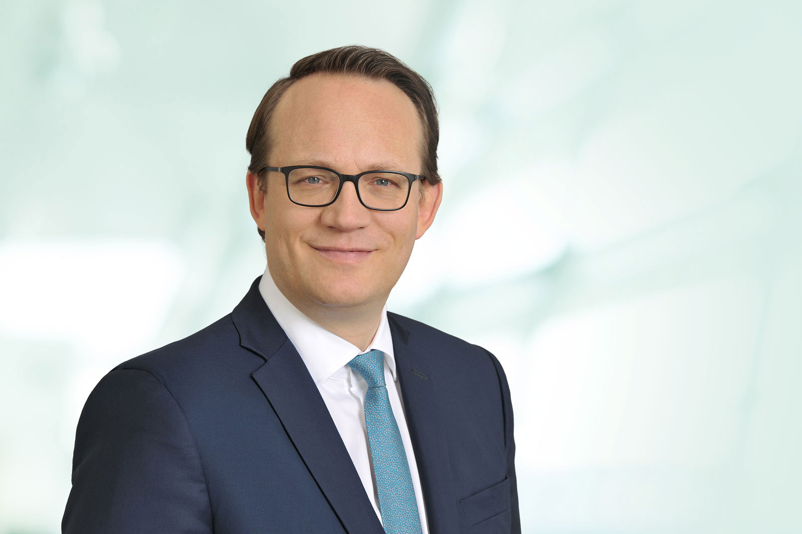 Ein Mann in einem Anzug mit Krawatte, der vor einem hellen, verschwommenen Hintergrund steht. Portrait von Dr. Markus Krebber.
