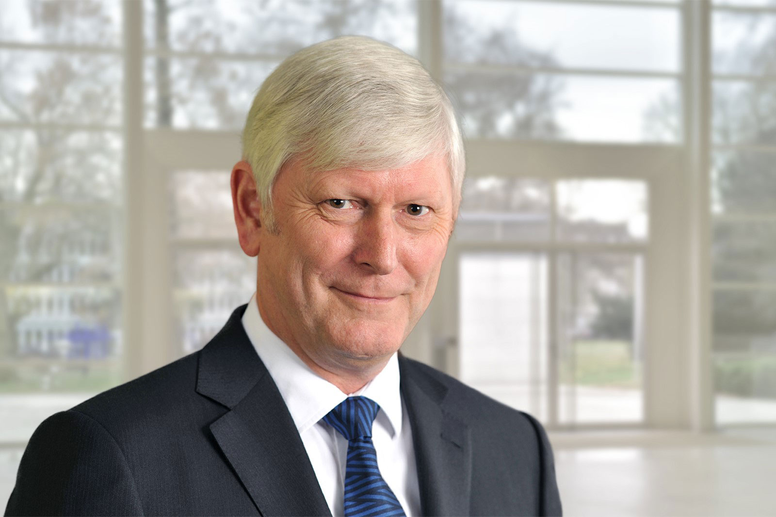 Ein Mann in einem dunkelblauen Anzug mit einer gestreiften Krawatte steht in einem modernen, hellen Raum. Portrait von Rolf Martin Schmitz.