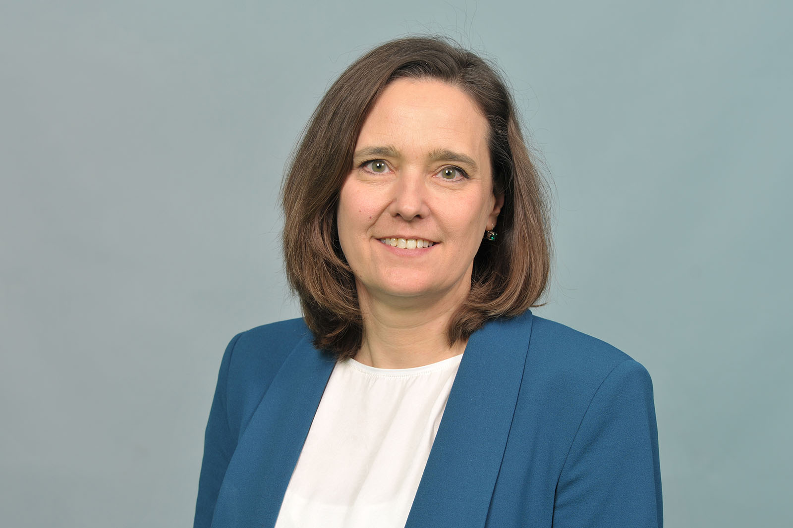 A woman with shoulder-length brown hair wears a blue blazer and a white blouse against a pale gray background. Portrait of Andrea Rodemann.