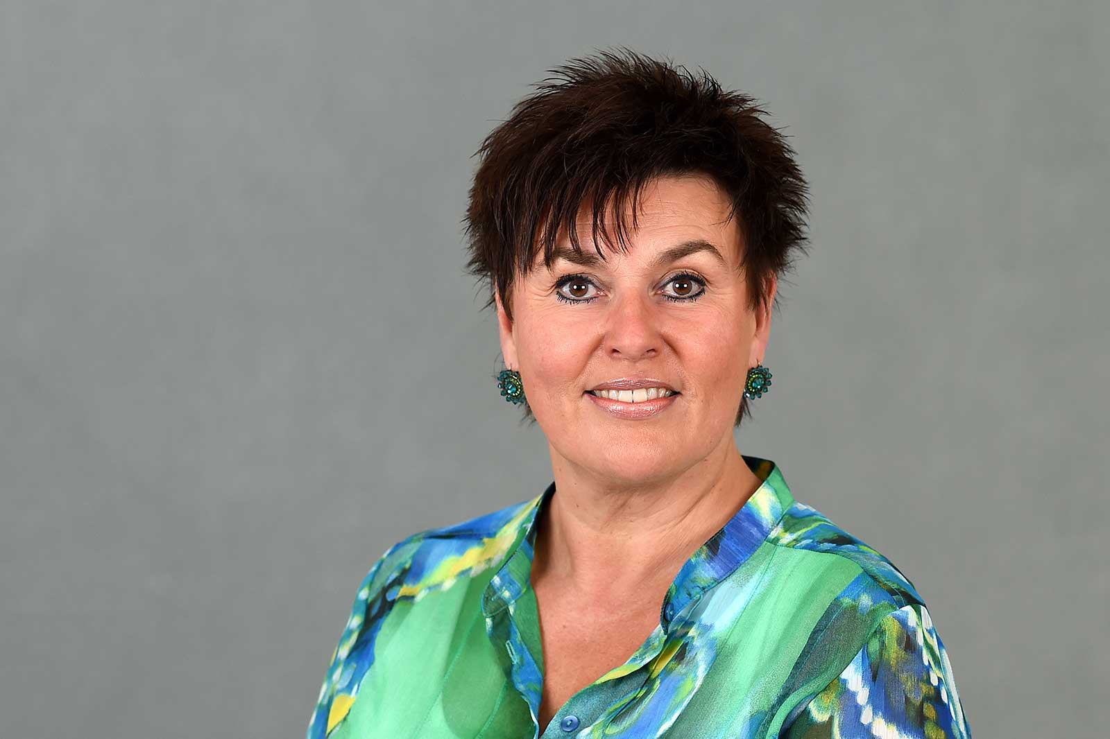Frau mit kurzem, dunklem Haar trägt ein buntes Hemd und grüne Ohrringe vor einem grauen Hintergrund. Portrait von Sandra Bossemeyer.