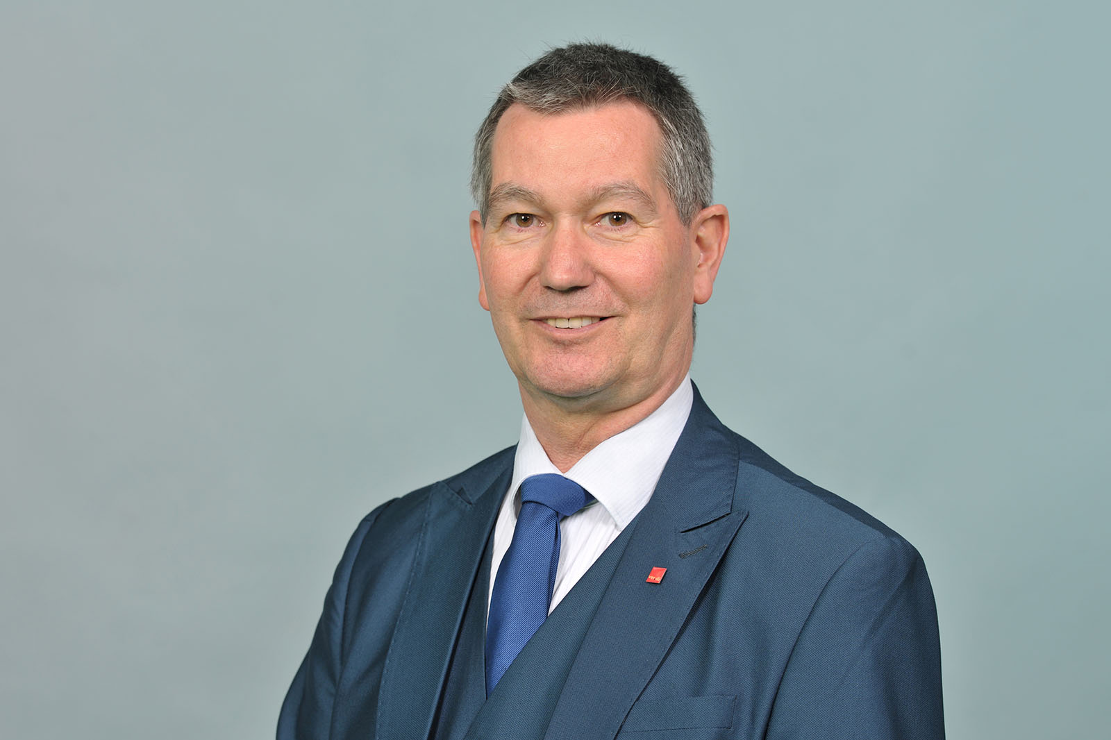 Eine Person mit kurzen grauen Haaren trägt einen dunkelblauen Anzug, weißes Hemd und blaue Krawatte vor einem hellblauen Hintergrund. Portrait von Christoph Schmitz-Dethlefsen.
