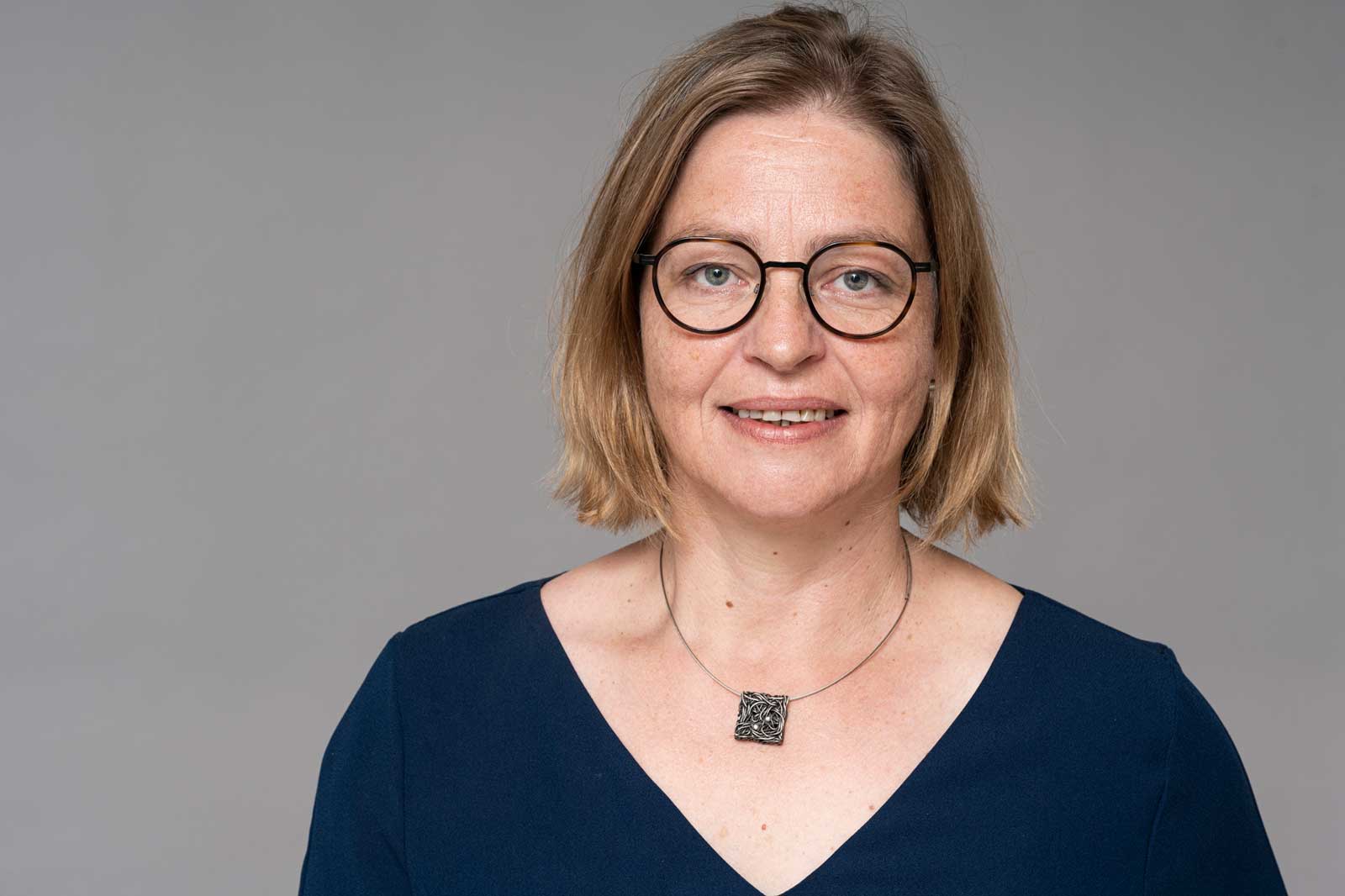 Die Person trägt ein dunkles Oberteil mit V-Ausschnitt und eine silberne Halskette mit einem quadratischen Anhänger. Portrait von Dagmar Paasch.