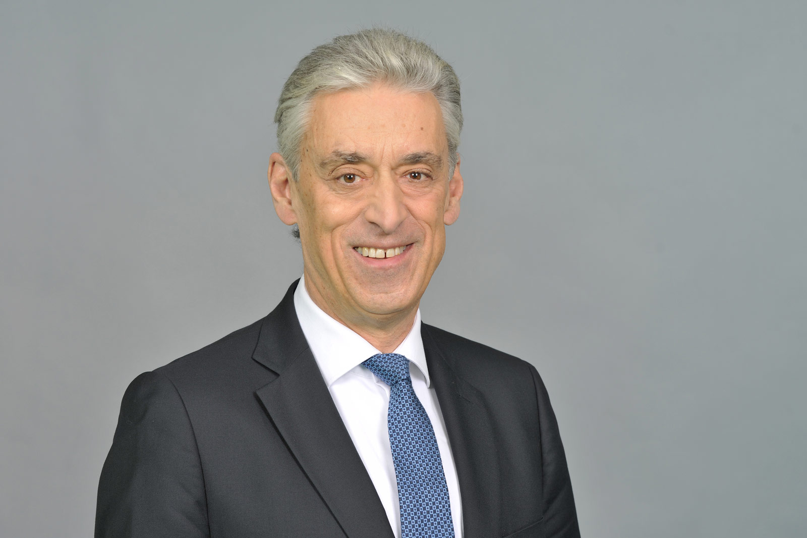Ein Mann in einem schwarzen Anzug mit einer blauen Krawatte vor einem grauen Hintergrund. Portrait von Dr. Frank Appel.