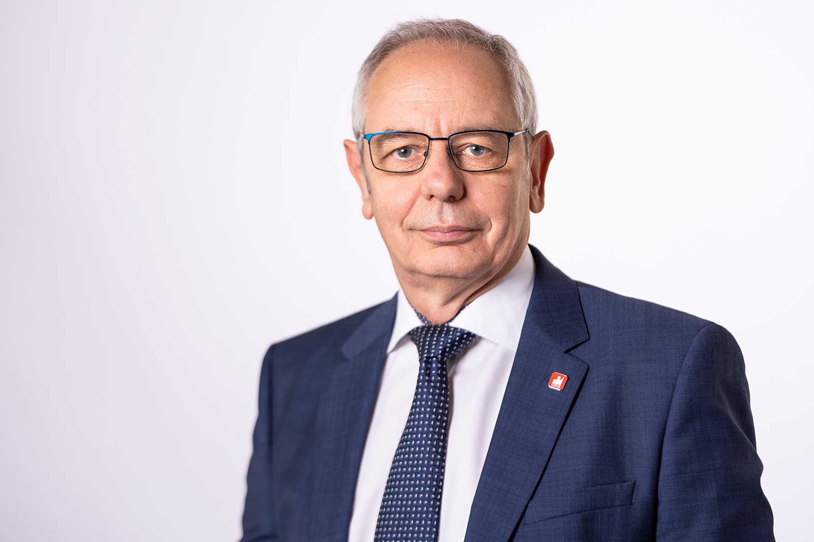 A man wearing a dark blue suit, a striped tie, and a red emblem on his lapel, set against a white background. Portrait of Michael Vassiliadis.