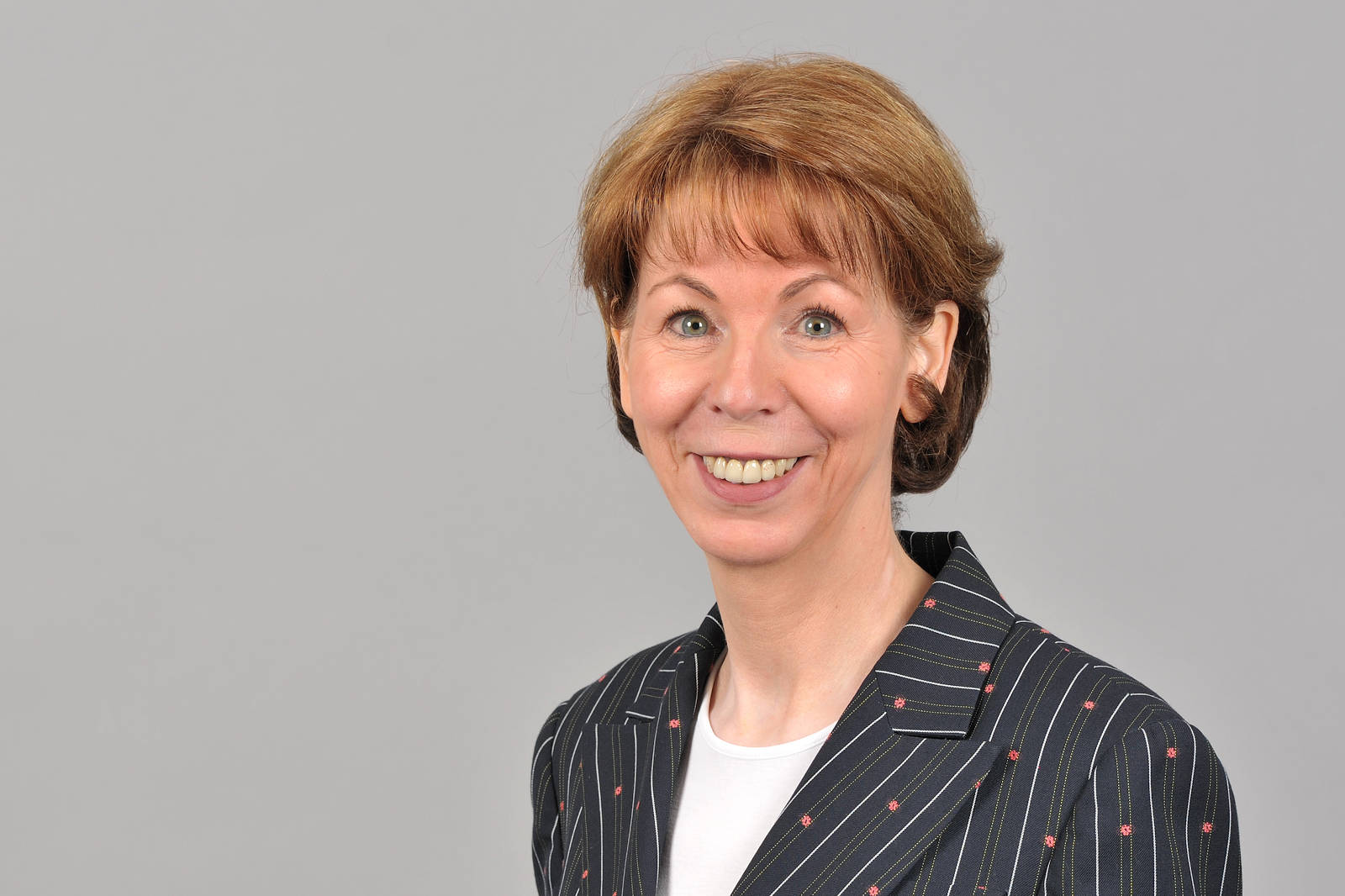 Eine Person mit braunem Haar trägt einen gestreiften Blazer und steht vor einem grauen Hintergrund. Portrait von Hauke Stars.