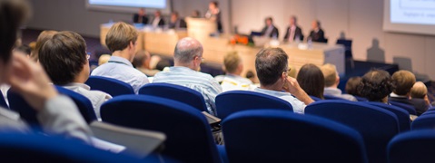 Zuhörer in einem Konferenzraum, die einem Vortrag von Rednern an einem Podium folgen.