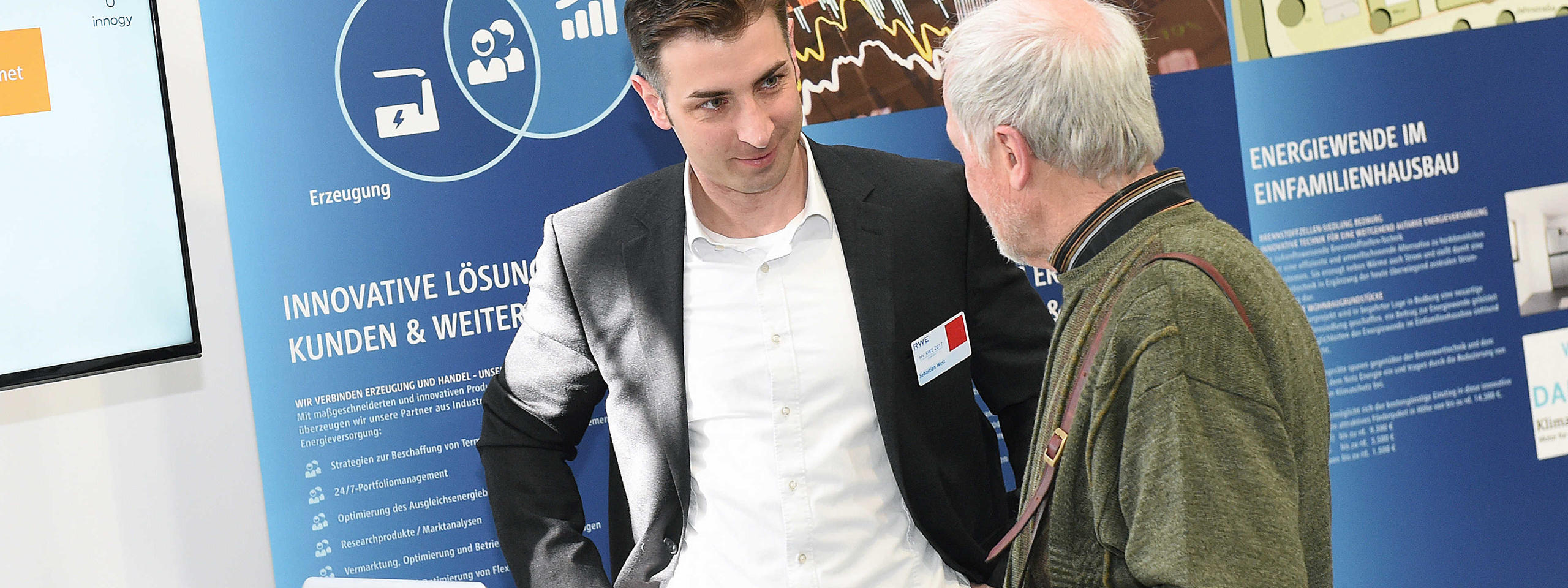Zwei Personen im Gespräch vor einem blauen Informationsstand über innovative Energielösungen. Ein Bildschirm ist sichtbar.