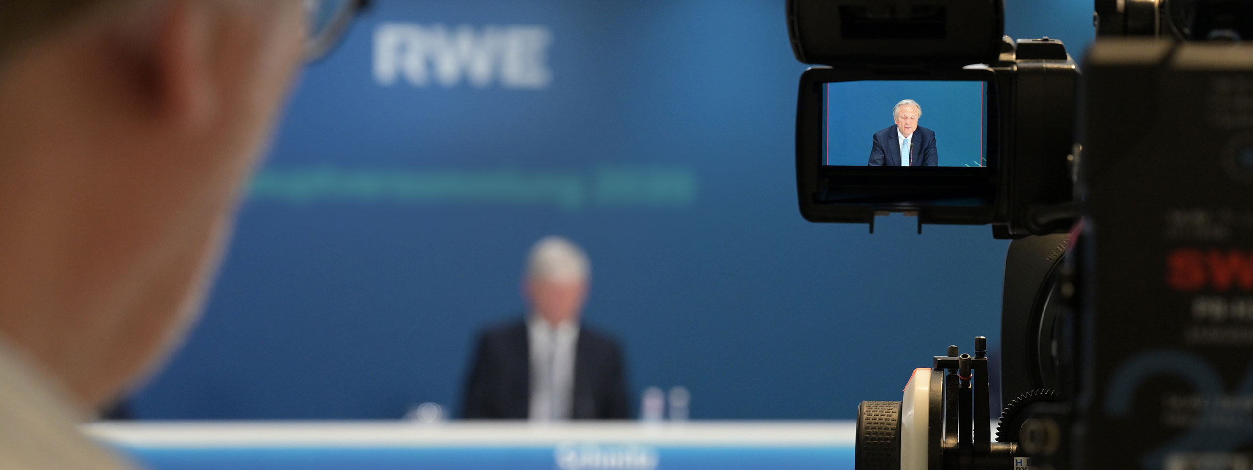 Eine Kamera filmt einen Redner bei einer Konferenz vor einem blauen Hintergrund mit dem RWE-Logo.