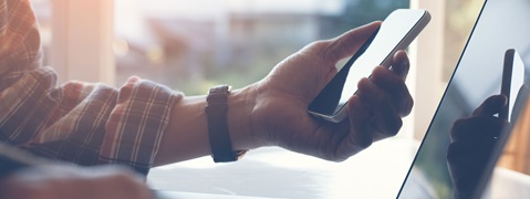Eine Hand hält ein Smartphone, während sie auf einen Laptop schaut. Ein Fenster im Hintergrund lässt Sonnenlicht durch.