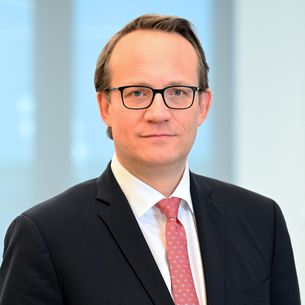 A man is wearing a suit with a red tie and white shirt. The background is bright and modern. Portrait of Dr Markus Krebber.