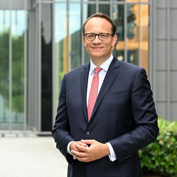 Ein Mann im Anzug steht vor einem modernen Gebäude. Er hat die Hände vor sich und trägt eine rote Krawatte. Portrait von Markus Krebber.