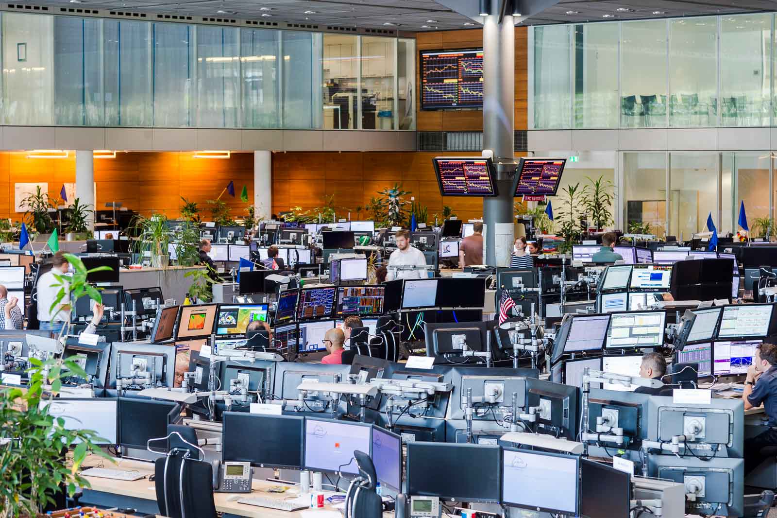 Ein modernes Büro mit vielen Arbeitsplätzen und Bildschirmen, wo Mitarbeiter aktiv arbeiten. Pflanzen sorgen für eine angenehme Atmosphäre.