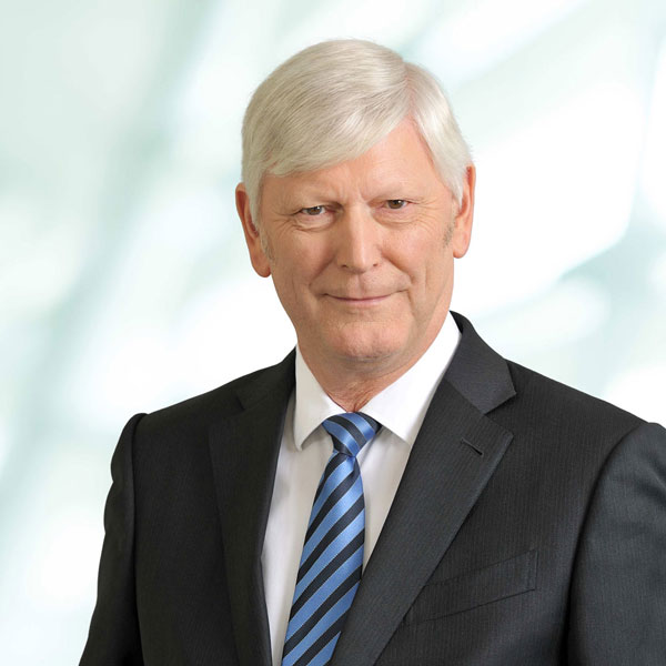 Ein Mann trägt einen schwarzen Anzug mit einer gestreiften Krawatte und hat graue Haare, vor einer hellen, unscharfen Hintergrund, Portrait von Dr. Rolf Martin Schmitz.