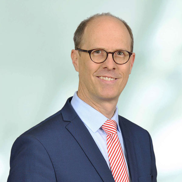 Ein Mann trägt einen Anzug mit gestreifter Krawatte und steht vor einem unscharfen Hintergrund, Portrait von Dr. Michael Müller.