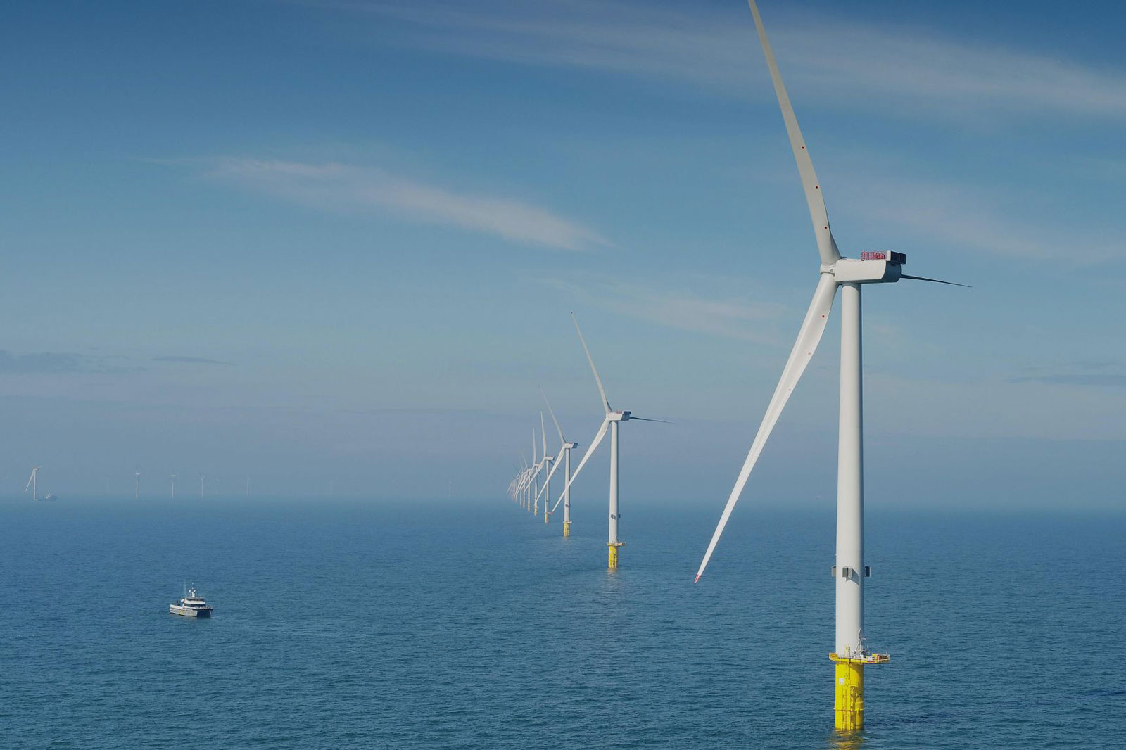 Eine Reihe von Offshore-Windkraftanlagen steht in einem ruhigen Meer unter einem klaren blauen Himmel, mit einem Boot in der Nähe.
