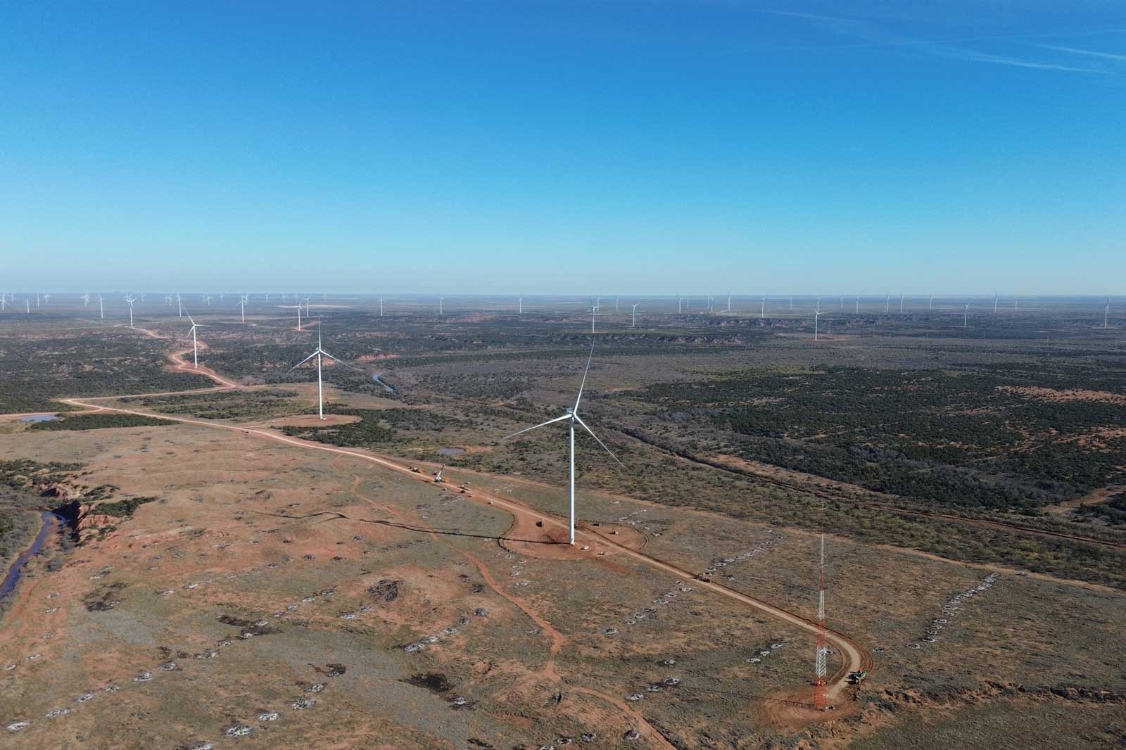 Luftaufnahme eines Windparks in einer offenen Landschaft mit Windkraftanlagen und einem blauen Himmel.