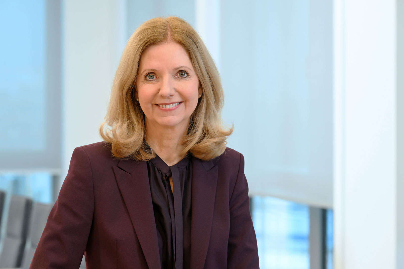 Eine Frau mit blonden, lockigen Haaren trägt einen dunklen Blazer und steht in einem modernen Büro. Portrait von Katja van Doren.