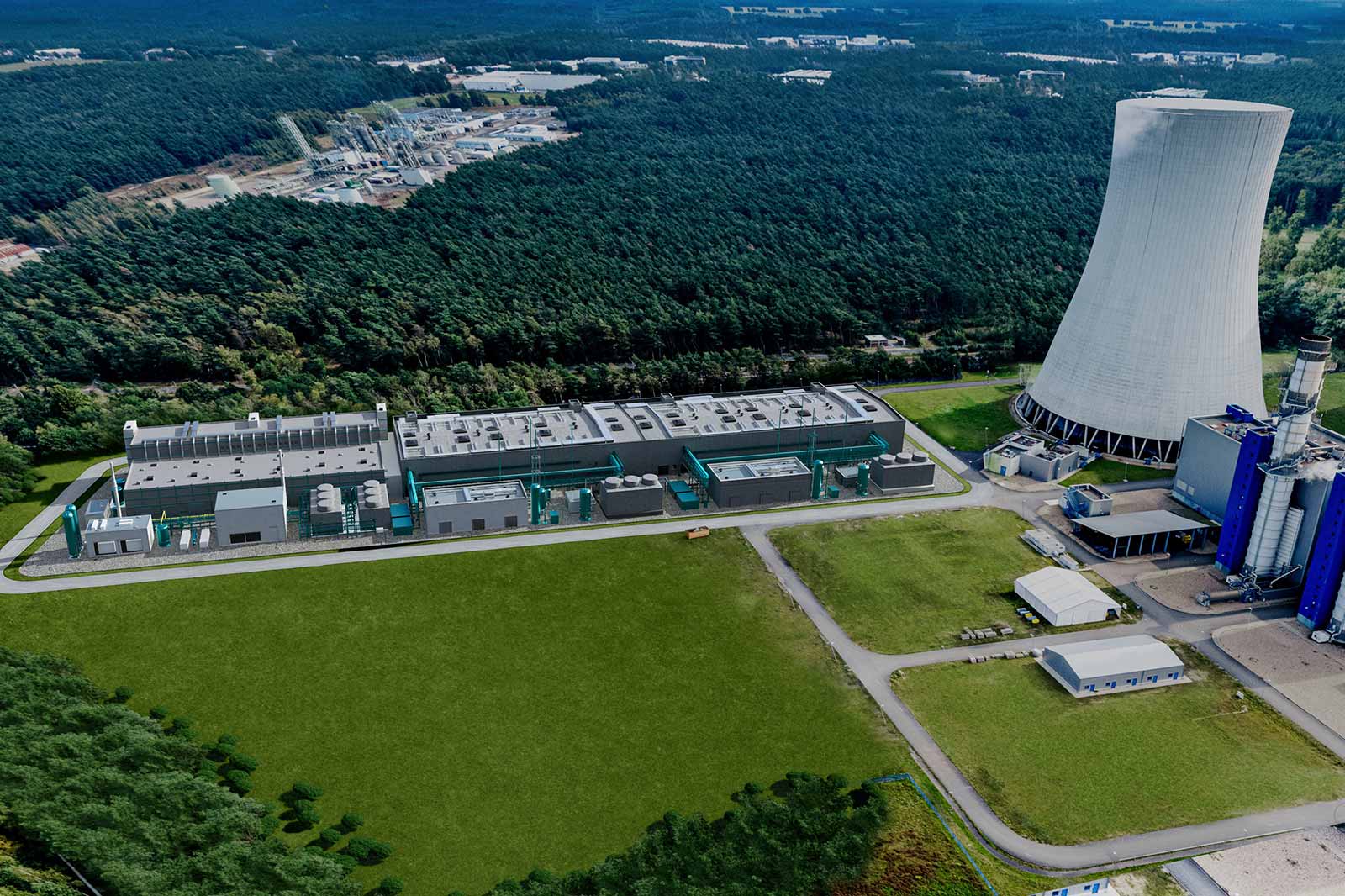 Luftaufnahme eines Industriekomplexes mit mehreren Gebäuden und einem Kühlturm, umgeben von Wald und Grünflächen.