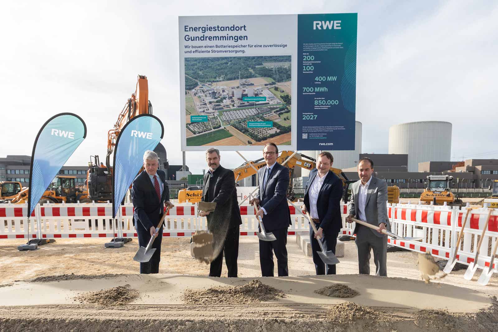 Fünf Männer in Anzügen stehen beim Spatenstich für einen neuen Energiestandort mit einem großen Informationsschild im Hintergrund.