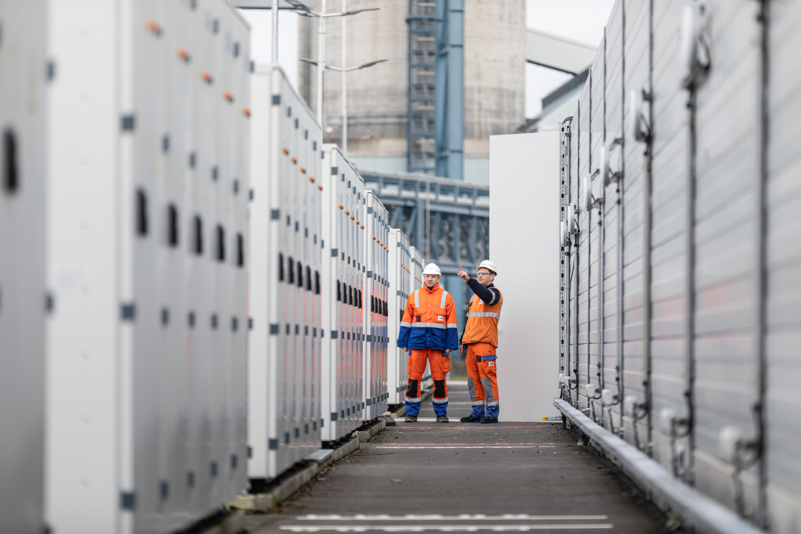 Two individuals in orange workwear stand between large electrical cabinets on an industrial site.