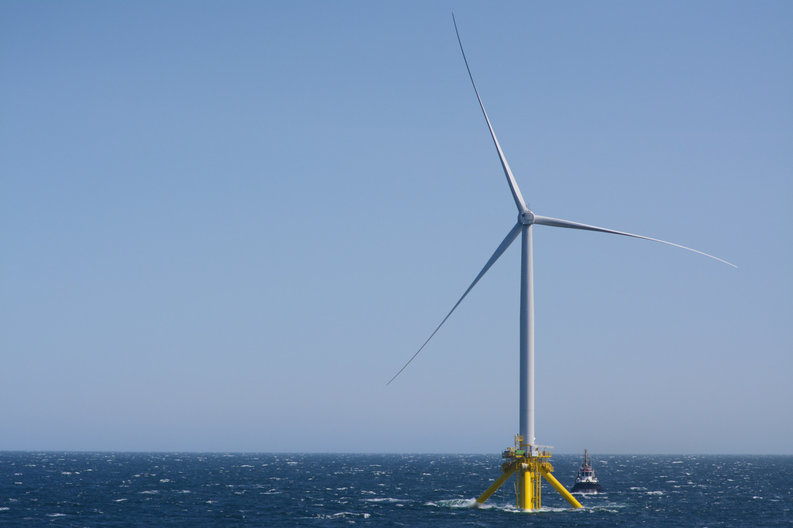 A large offshore wind turbine stands tall against a clear sky, with waves in the ocean and a small boat nearby.