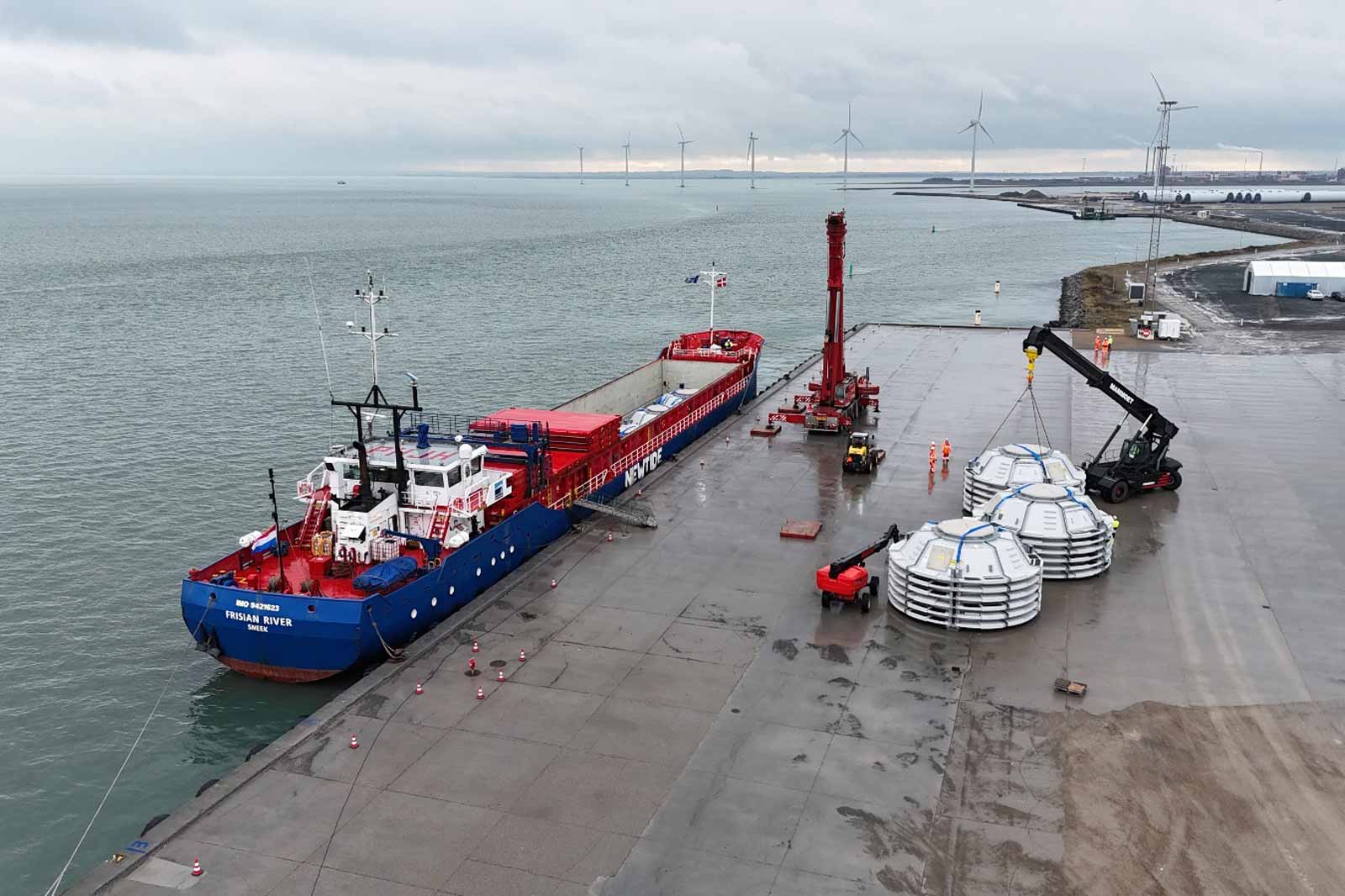 Ein Frachtschiff namens Frisian River liegt am Kai. Im Hintergrund sind Windturbinen zu sehen und Arbeiter laden große Materialien.