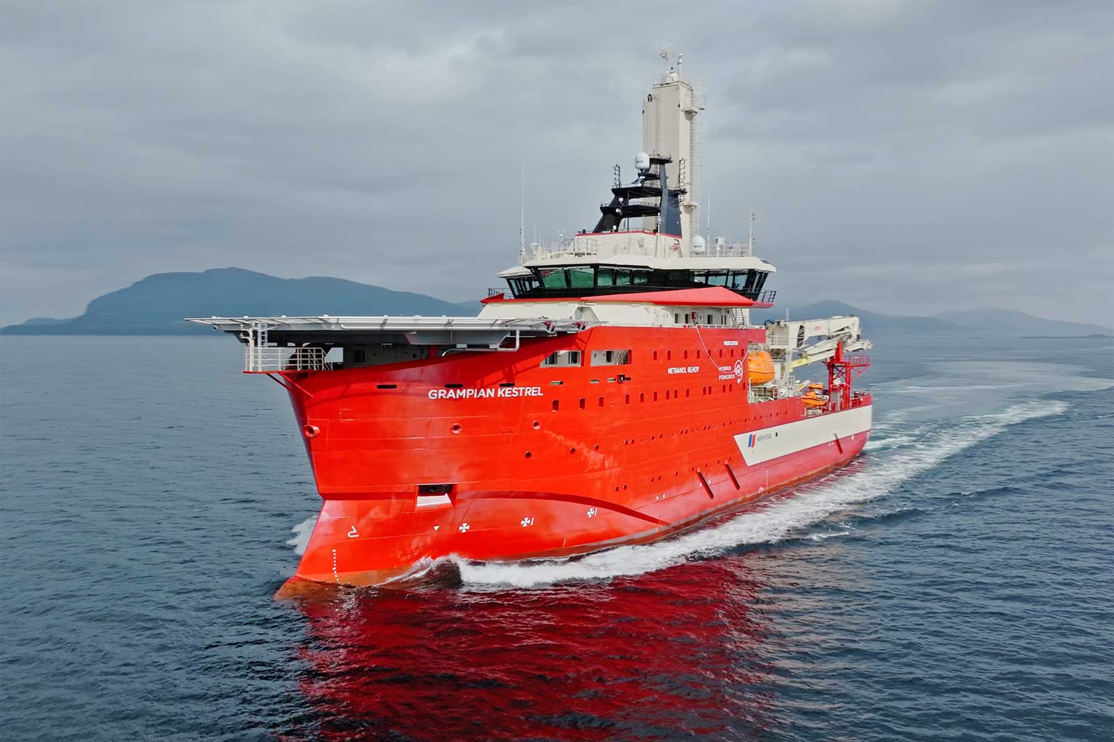 Ein großes rotes Schiff namens 'Grampian Kestrel', das ruhige Gewässer in der Nähe von Bergen unter einem bewölkten Himmel befährt.