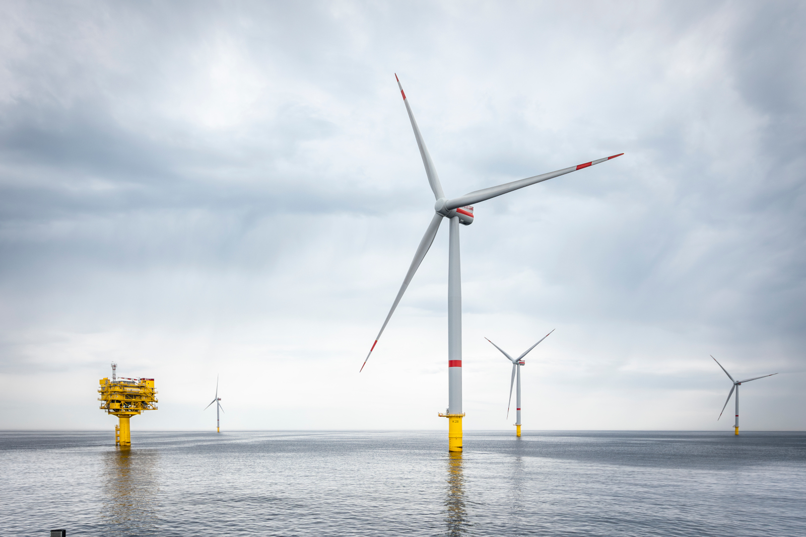 Mehrere Windkraftanlagen stehen im Wasser, mit grauem Himmel und reflektierender Wasseroberfläche. Eine Plattform ist sichtbar.