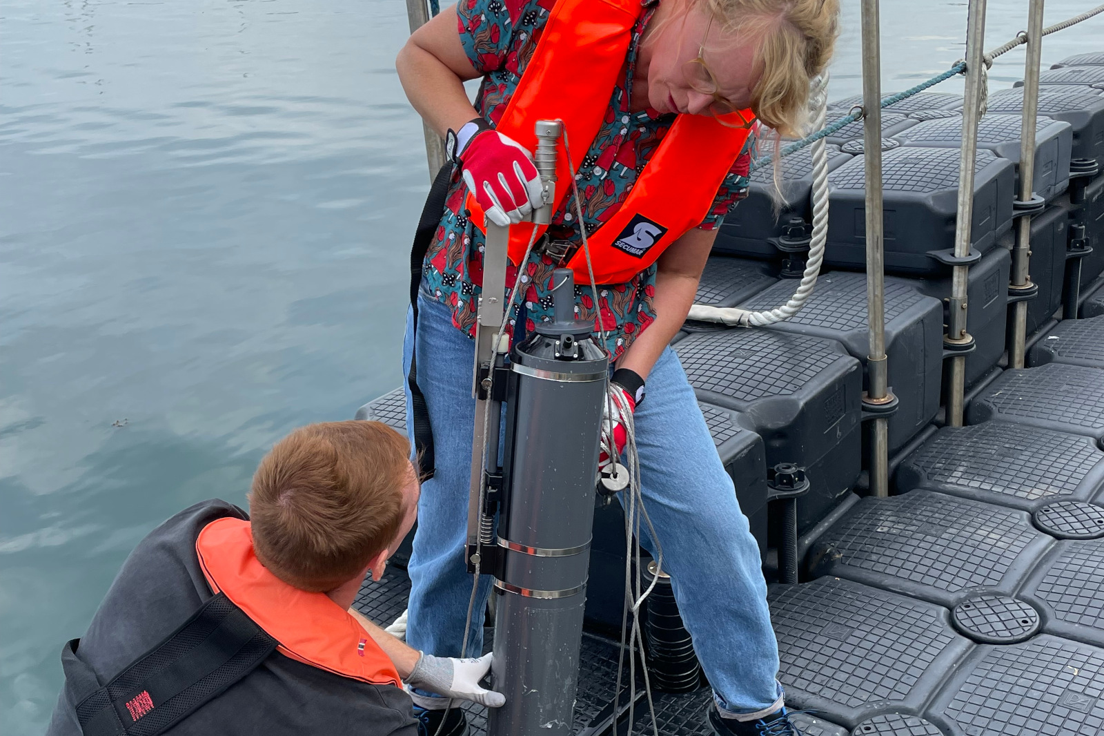 Eine Person in einem orangefarbenen Schwimmwesten hält ein Gerät auf einem Boot. Eine andere Person beobachtet die Handlung.