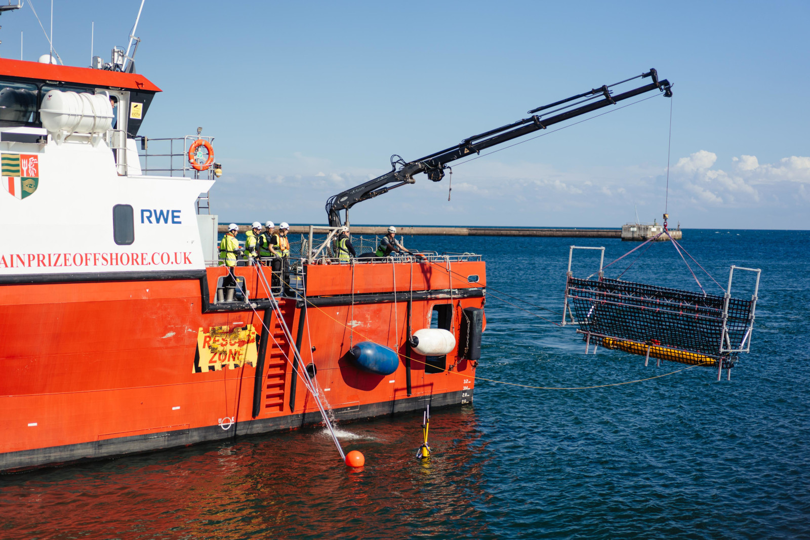 Ein orangefarbenes Schiff hebt eine schwere Last mit einem Kran über dem Wasser. Arbeiter in Schutzkleidung stehen in der Nähe.