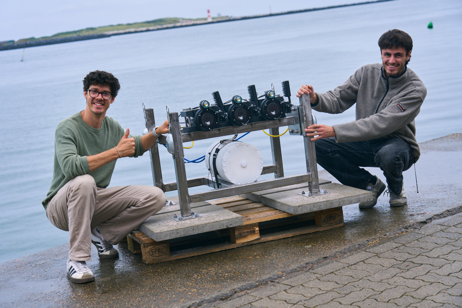Zwei Personen präsentieren ein technisches Gerät mit Kameras, das an einem Wasserweg platziert ist.