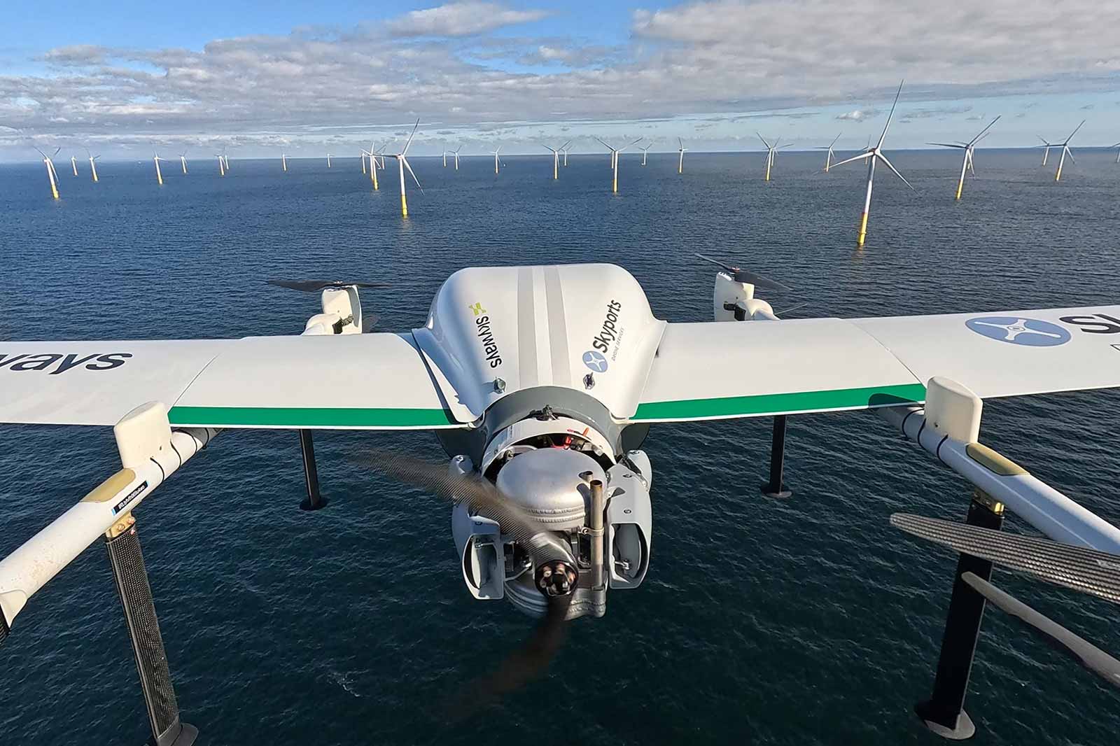 Eine Drohne schwebt über dem Wasser mit Windkraftanlagen im Hintergrund, die in einer Vielzahl angeordnet sind.