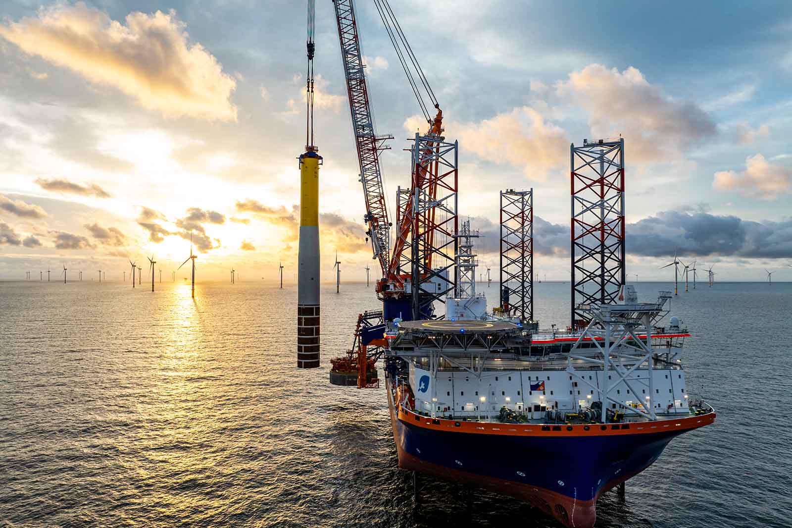 Ein Offshore-Baukran hebt einen großen Windkraftturm bei Sonnenuntergang, neben einem großen Arbeitsschiff mit vier Pfeilern im Meer.
