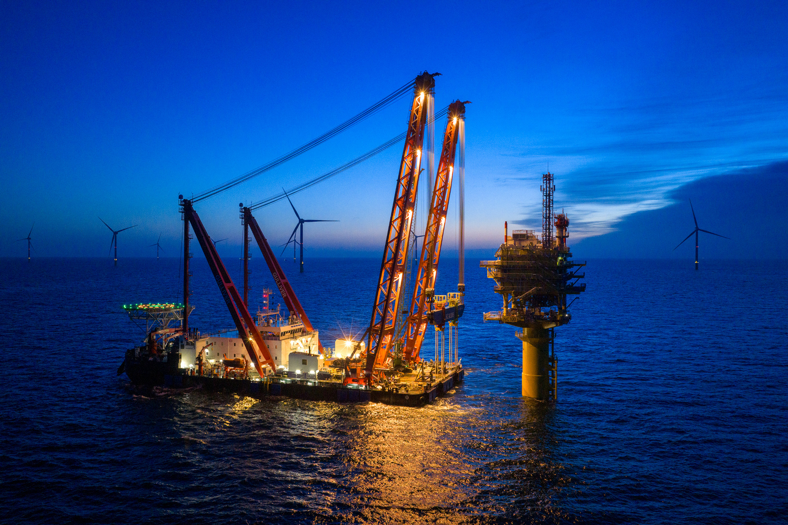 Offshore heavy lift vessel with cranes beside lit platform and wind turbines.