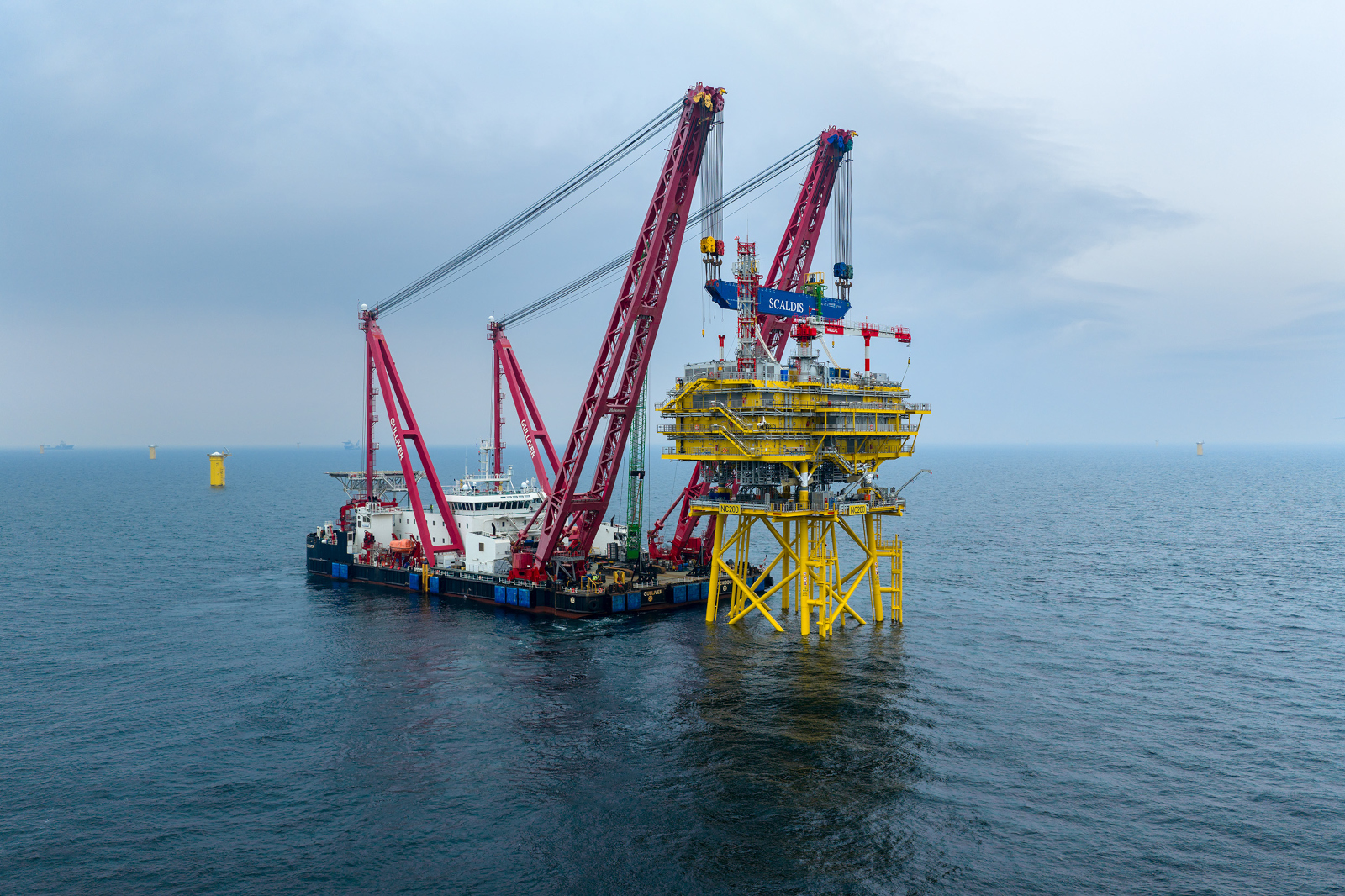 Heavy lift vessel with red cranes installing yellow offshore platform