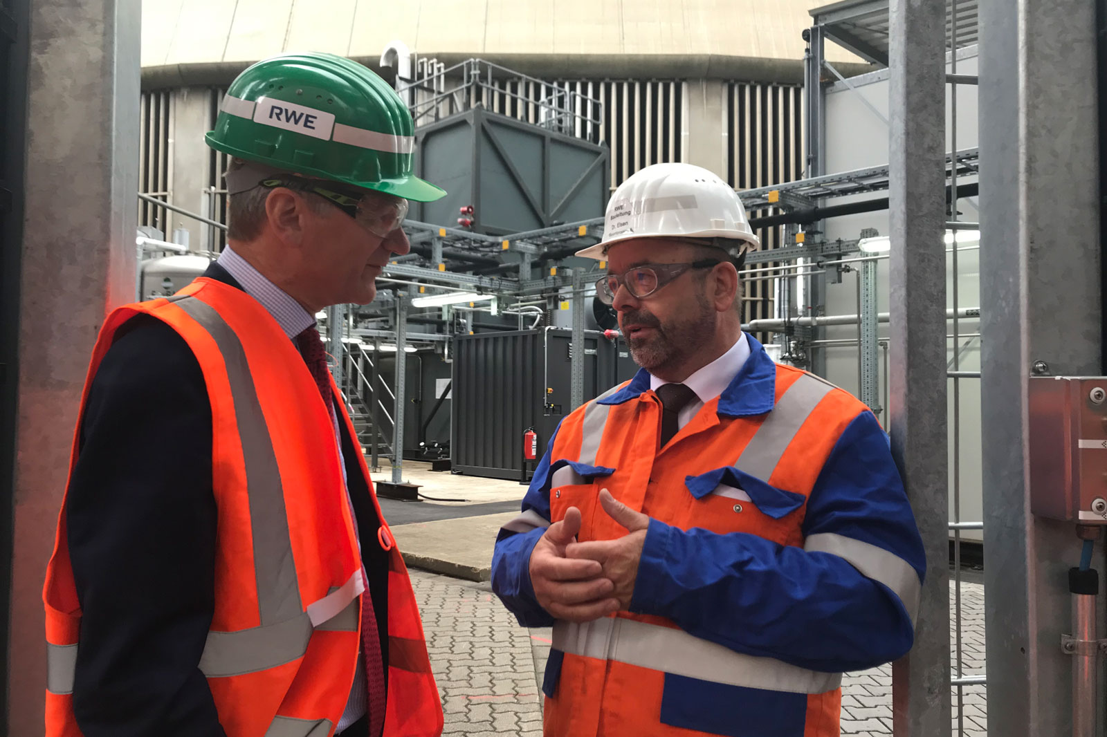 Zwei Personen in Sicherheitskleidung diskutieren in einem Industriegebäude, während im Hintergrund Maschinen sichtbar sind.