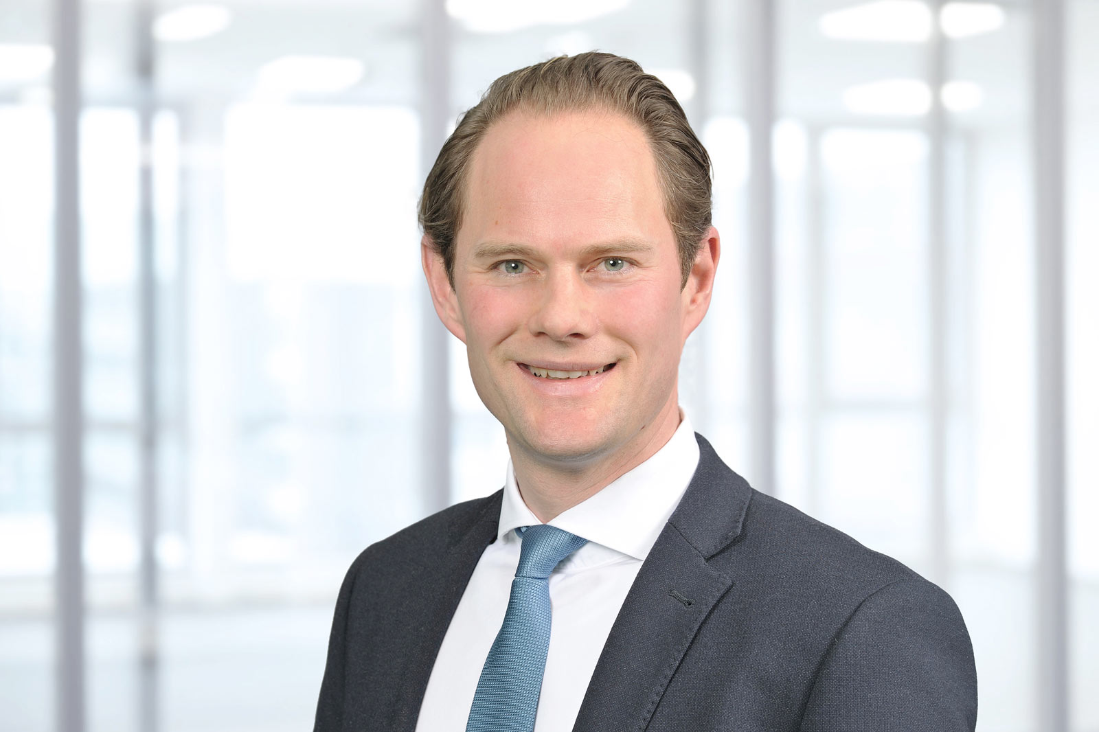 Ein Mann in einem eleganten Anzug und einer Krawatte steht vor einem verschwommenen Büro-Hintergrund und strahlt Professionalität aus.